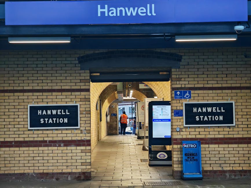 A view of Hanwell Station entrance with two black rectangular signs displaying 'HANWELL STATION' mounted on a brick wall. The station features a brick archway leading through to the platform area, whe
